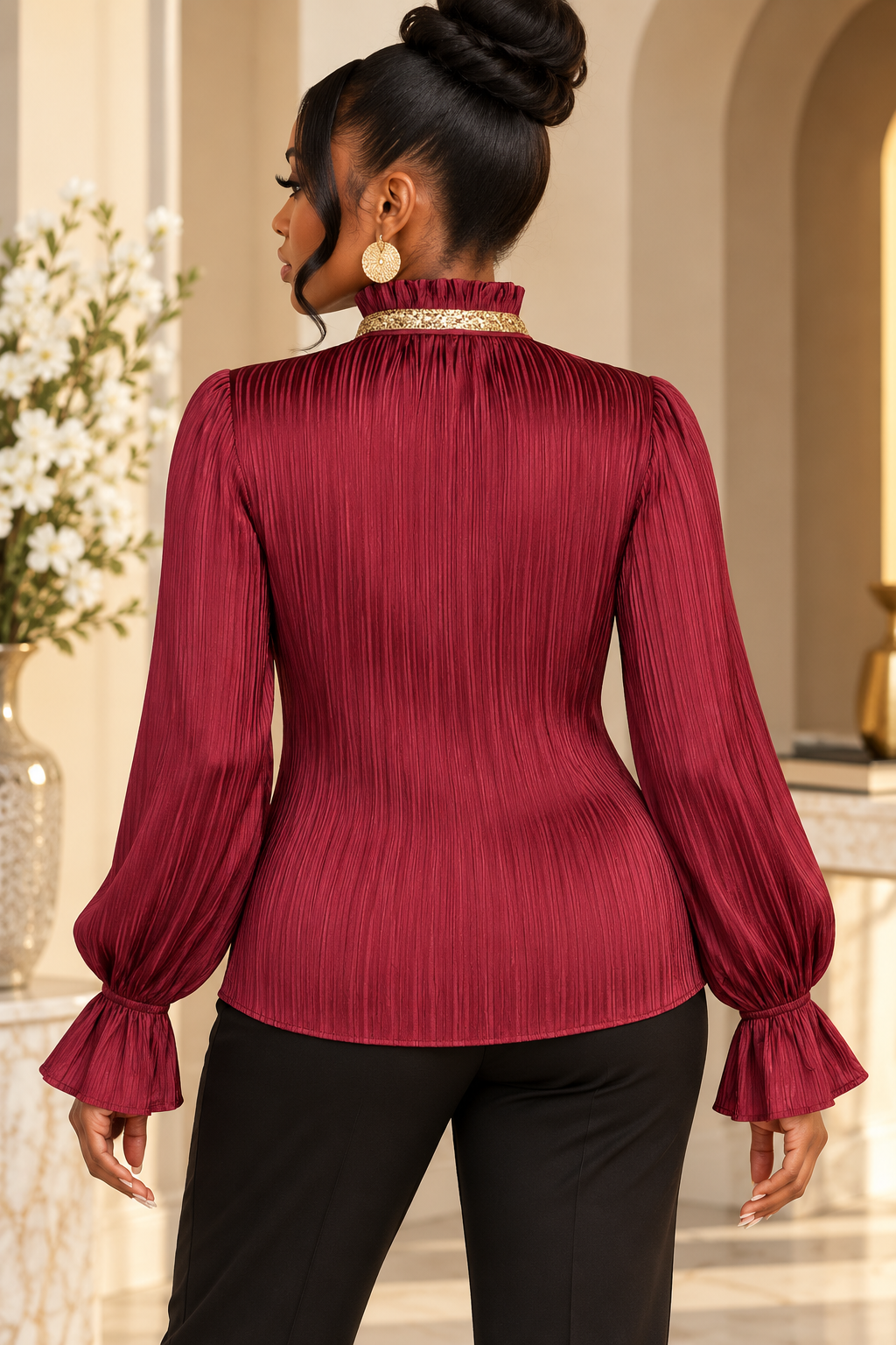 A woman shown from the back wearing a pleated long-sleeve blouse in deep burgundy with a high ruffled collar and flared cuffs, paired with black pants in an indoor setting.