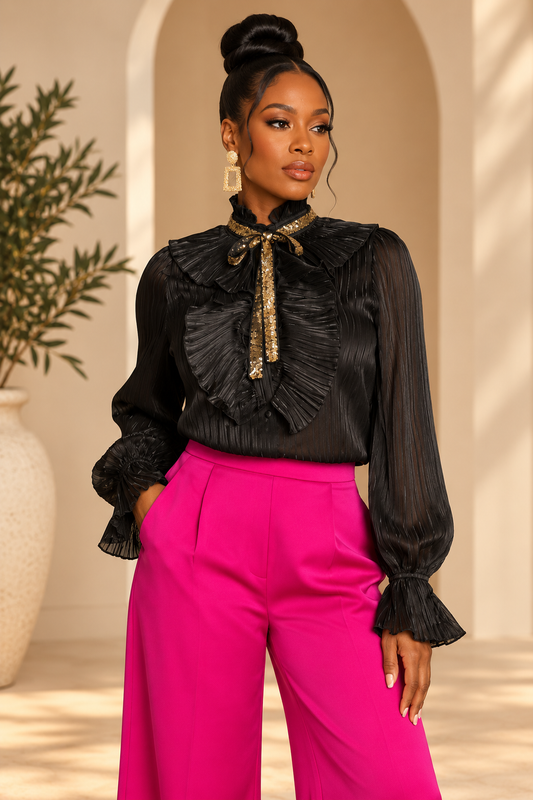 A woman modeling a black pleated long-sleeve blouse with a ruffled high collar and a gold patterned bow tie detail, paired with bright pink trousers, in a warm-toned indoor setting with arches.