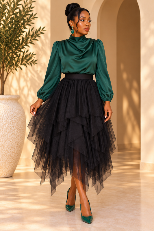 A woman models a two-piece outfit featuring a dark green satin mock-neck top with puffed sleeves and a layered black tulle high-low skirt, paired with green pointed-toe heels in a sunlit architectural setting.