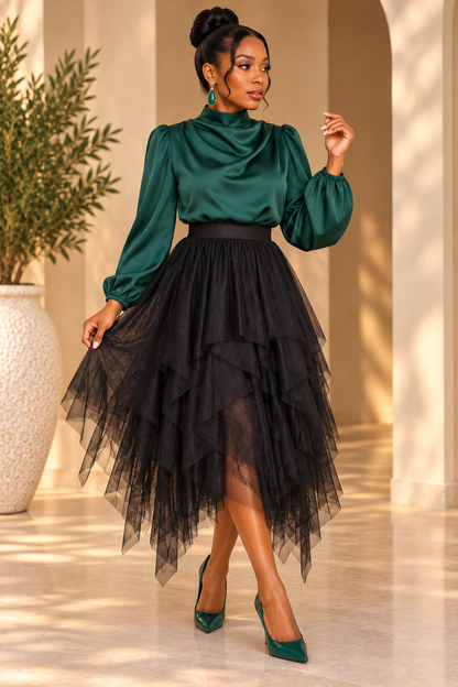 A woman models a two-piece outfit featuring a dark green satin long-sleeve mock-neck top and a black tiered tulle midi skirt, paired with matching green pointed-toe heels, set against a warm beige architectural background.