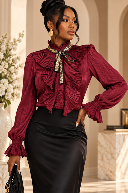 A woman models a long-sleeve burgundy pleated blouse with a high ruffled collar and a jeweled bow tie detail, paired with a black satin pencil skirt in an elegant indoor setting.